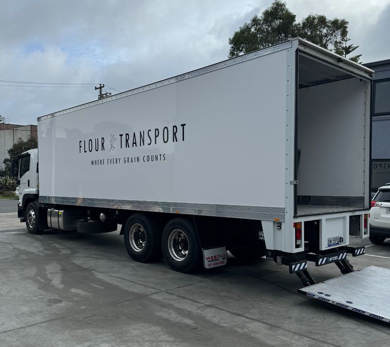 The Flour Transport delivery truck, signwritten 'Flour Transport – Where every grain counts', with its tailgate lift lowered